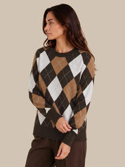 A woman wearing a long-sleeve argyle sweater in brown, tan, white, and dark brown, paired with dark brown pants, set against a solid beige background.