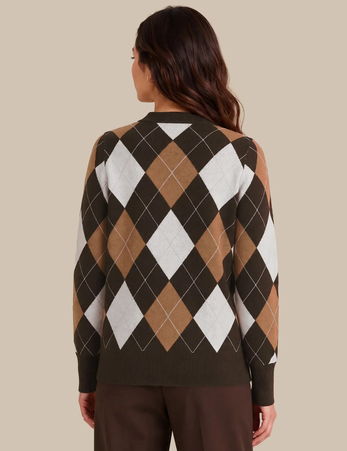 A woman viewed from the back wearing a long-sleeve argyle sweater with dark brown, tan, white, and black diamonds, paired with dark brown pants, against a solid beige background.