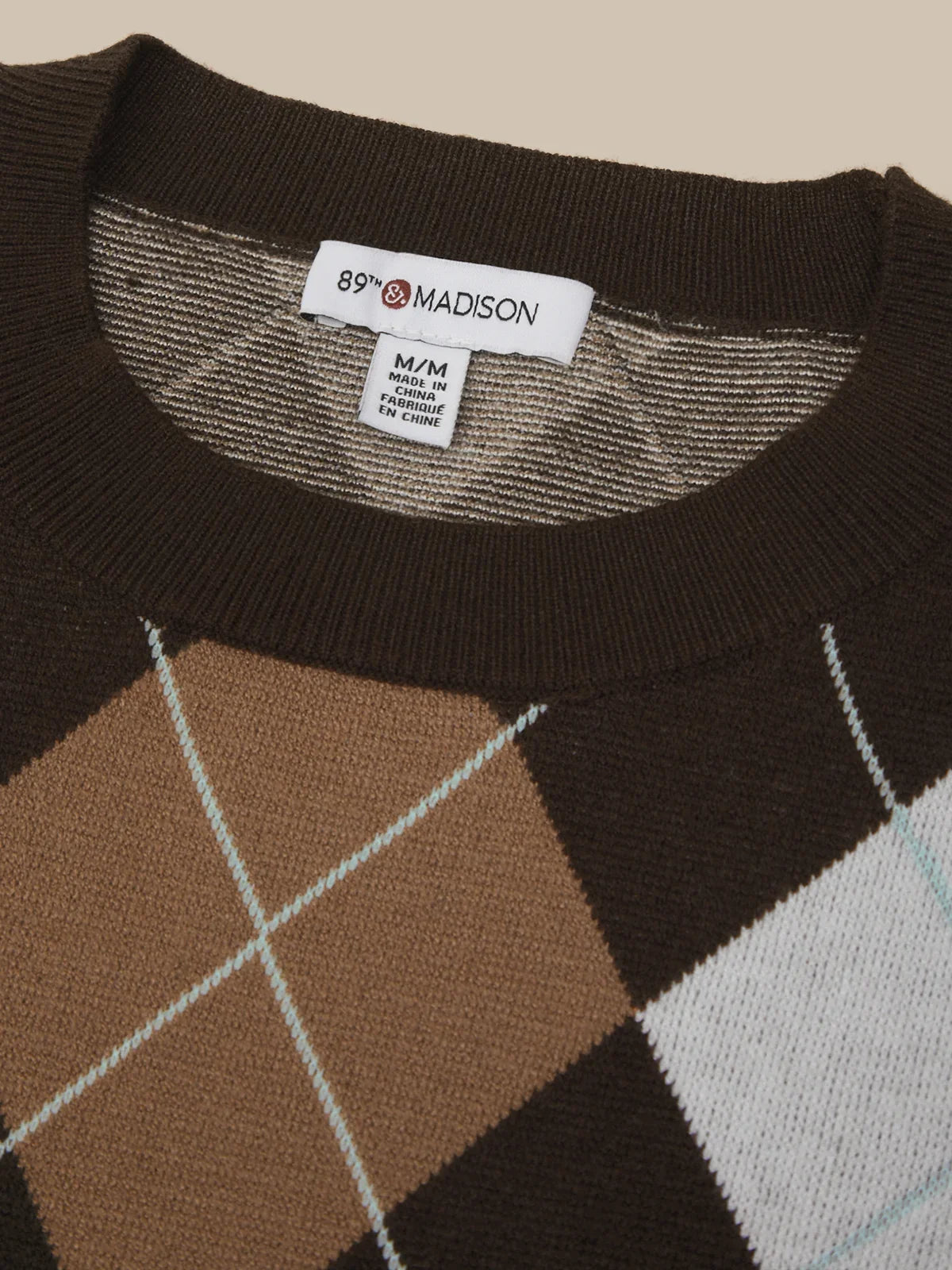 Close-up of a knit sweater neck area showing a white label that reads 89% MADISON. The sweater has a dark brown ribbed collar and upper section with an argyle pattern in brown, tan, and white.