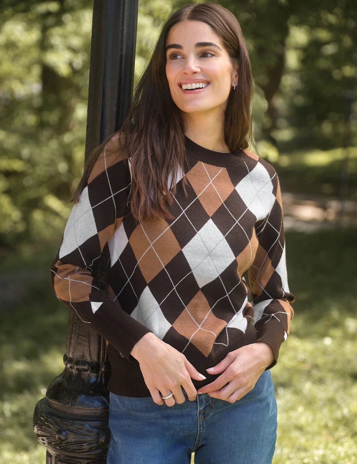 A woman wearing a brown, tan, and white argyle sweater leans against a dark metal pole in a sunny park setting.