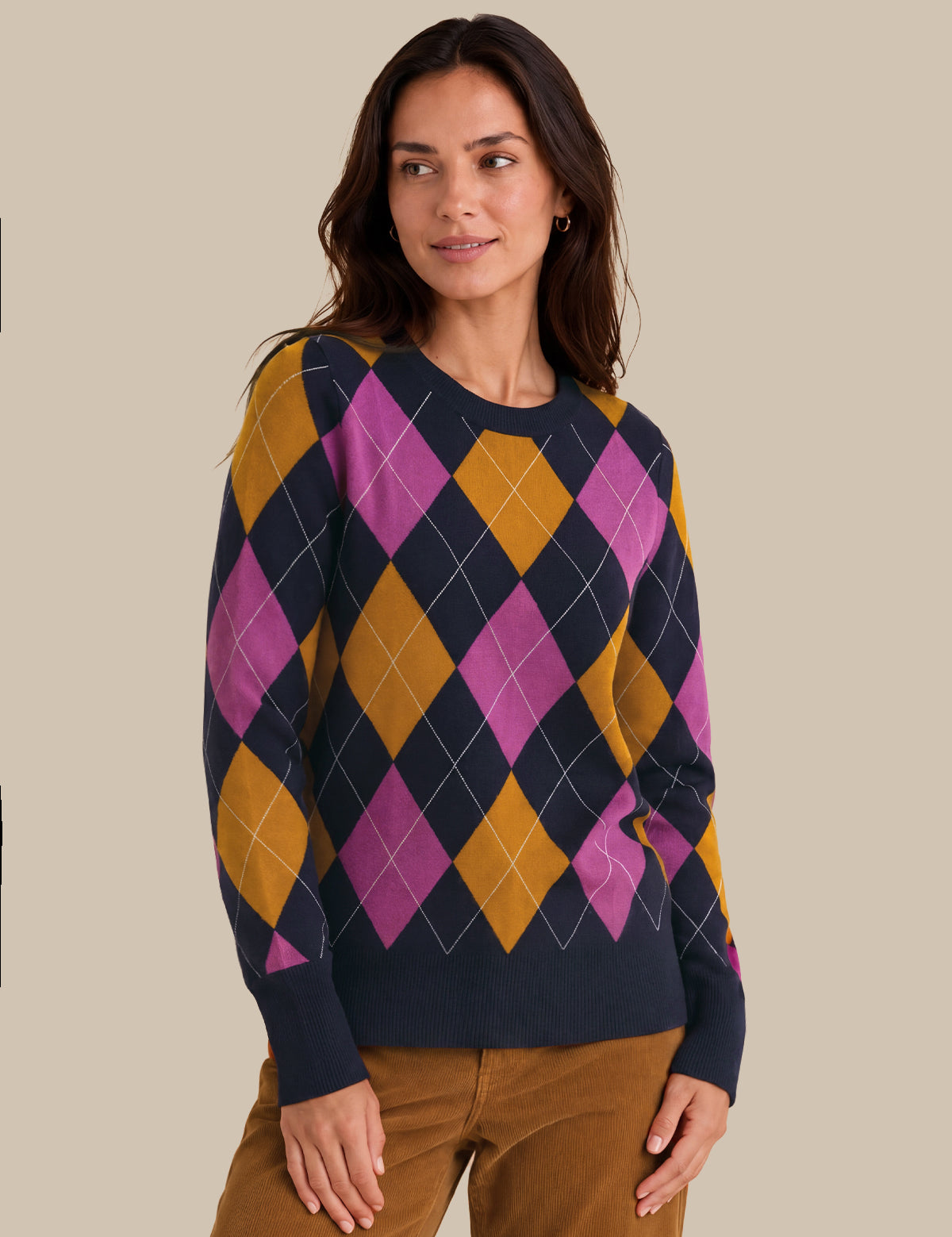 A woman wearing a navy blue crewneck sweater with a vibrant argyle pattern in magenta pink and mustard yellow, paired with brown pants, set against a solid beige background.