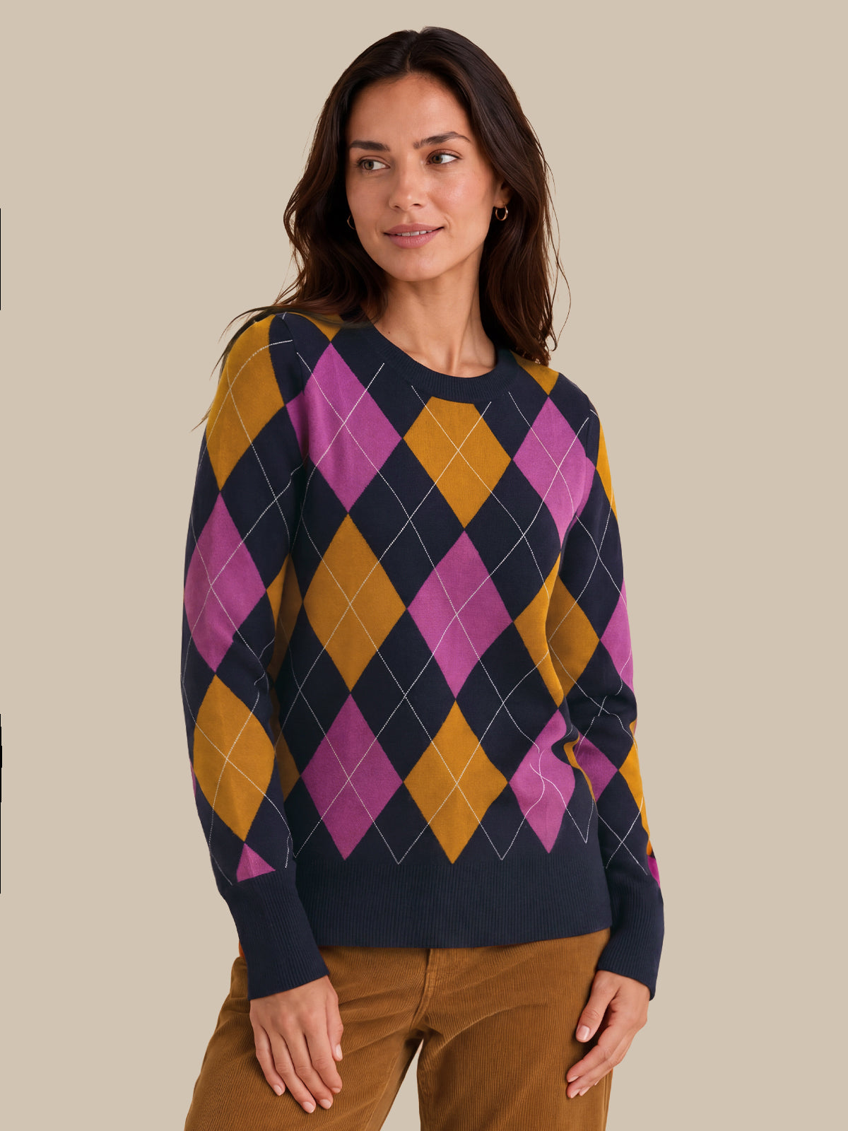A woman wearing a navy blue crewneck sweater with a vibrant argyle pattern in magenta pink and mustard yellow, paired with brown pants, set against a solid beige background.