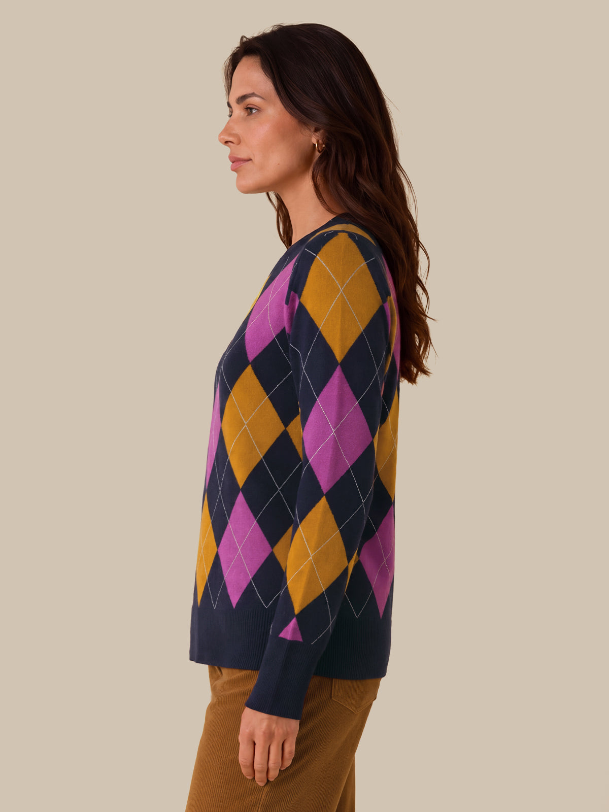 A woman shown in side profile wearing a long-sleeve knit sweater with a large argyle pattern in navy, mustard yellow, and magenta, paired with brown pants, against a solid beige background.