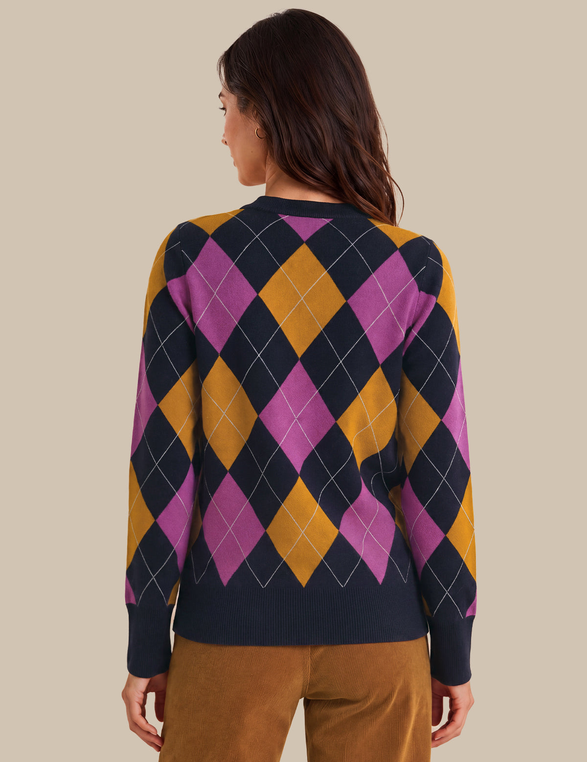 A woman shown from the back wearing a long-sleeve argyle sweater with navy, magenta, and mustard yellow diamonds, paired with brown pants, against a solid beige background.