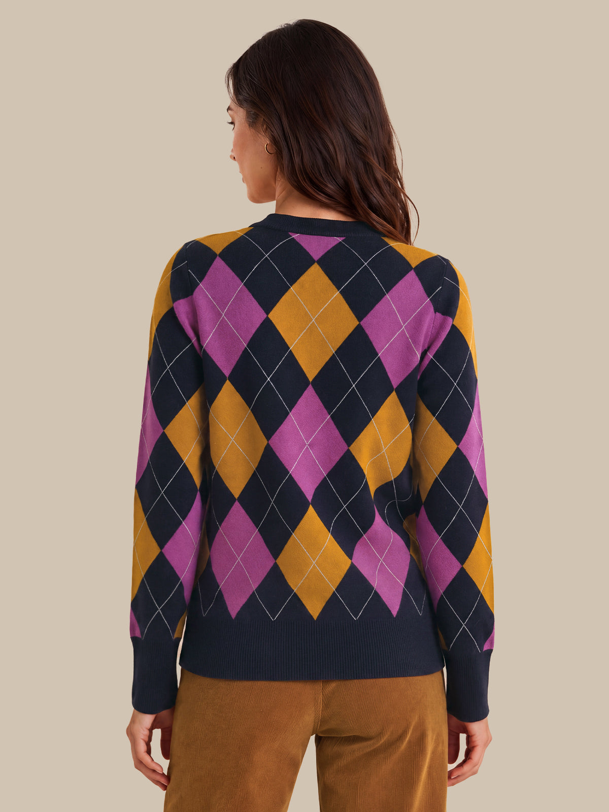 A woman shown from the back wearing a long-sleeve argyle sweater with navy, magenta, and mustard yellow diamonds, paired with brown pants, against a solid beige background.