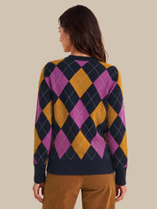 A woman shown from the back wearing a long-sleeve argyle sweater with navy, magenta, and mustard yellow diamonds, paired with brown pants, against a solid beige background.
