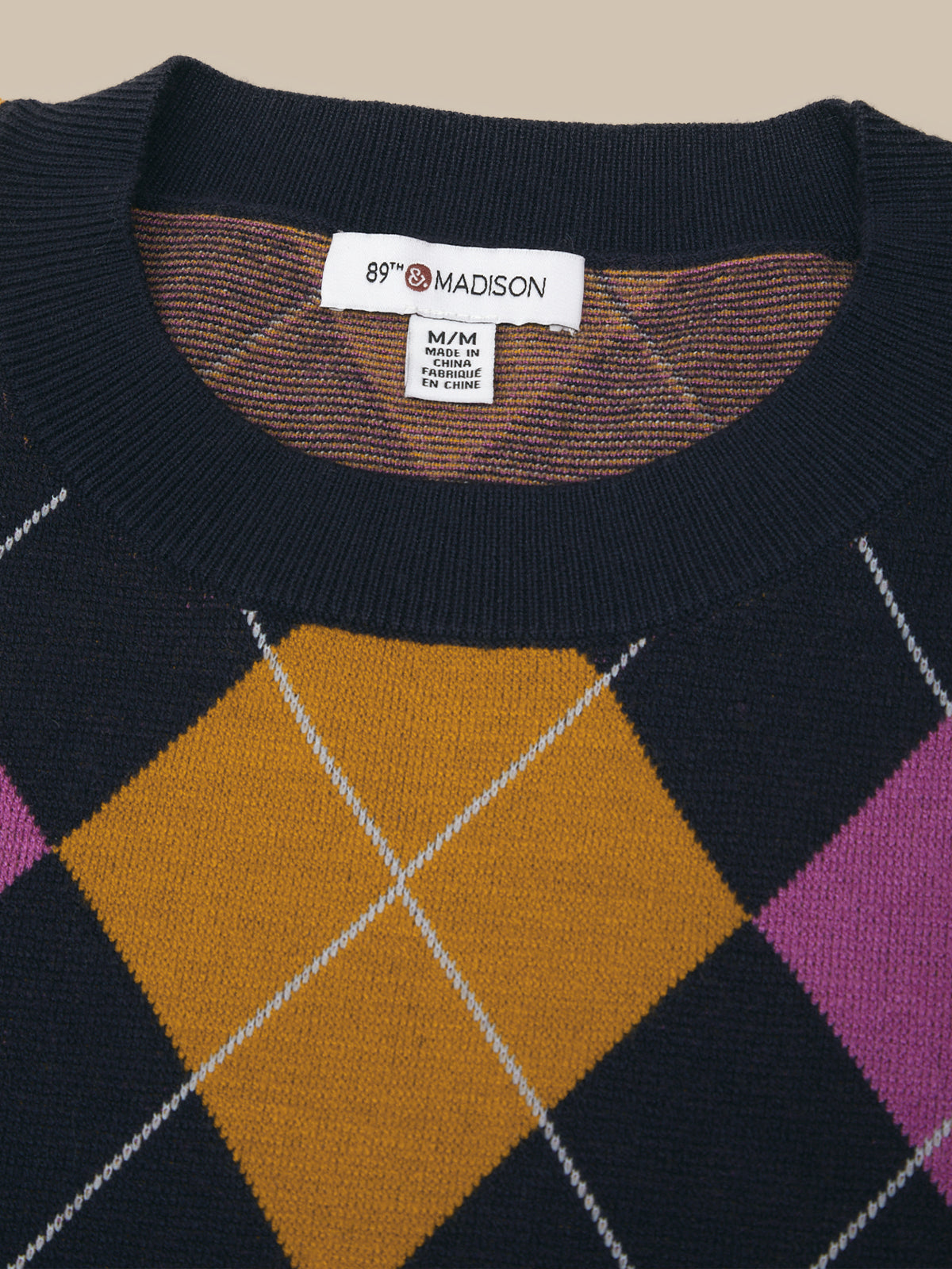 Close-up of a knit sweater with a navy base and an argyle pattern in mustard yellow, purple, and brown, featuring a visible Madison brand label at the collar.