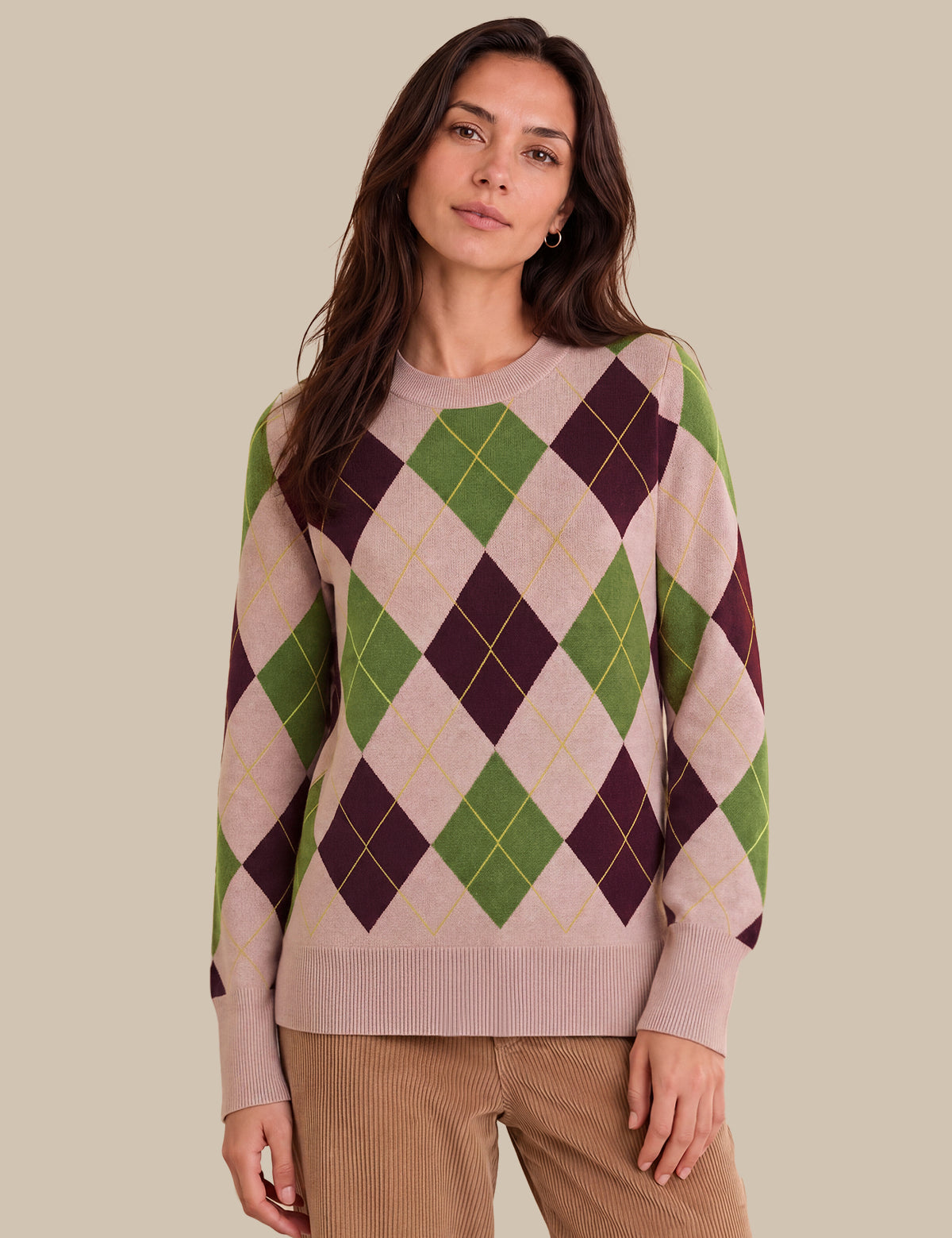 A woman wearing a long-sleeve argyle sweater in beige, green, and dark purple, paired with brown corduroy pants, against a solid beige background.