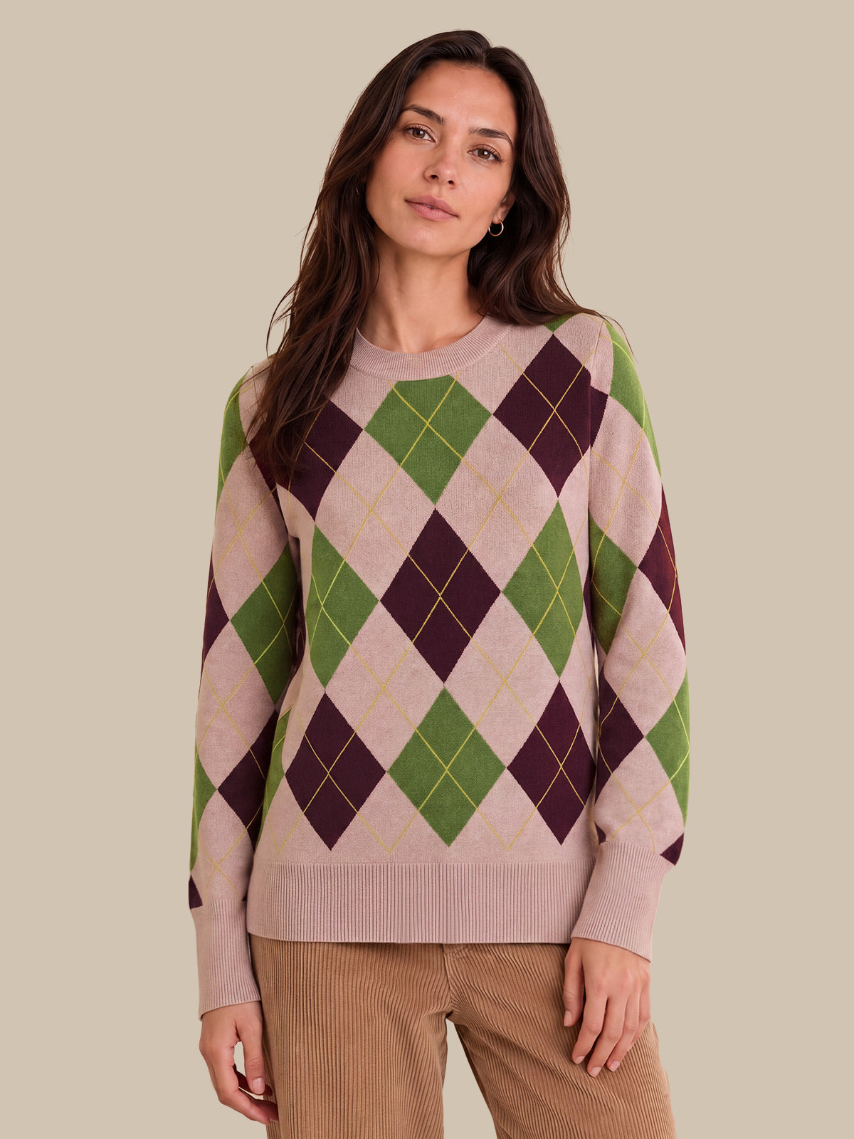 A woman wearing a long-sleeve argyle sweater in beige, green, and dark purple, paired with brown corduroy pants, against a solid beige background.