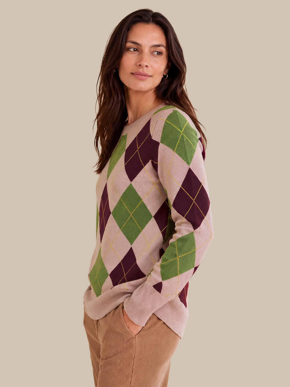A woman wearing a long-sleeve argyle sweater with green, burgundy, and beige diamonds, paired with tan corduroy pants, set against a solid beige background.