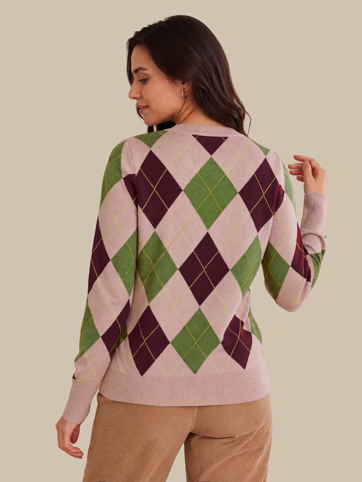 A woman shown from the back wearing a long-sleeve knit sweater with an argyle pattern in light pink, green, and dark purple, paired with tan pants, against a solid beige background.