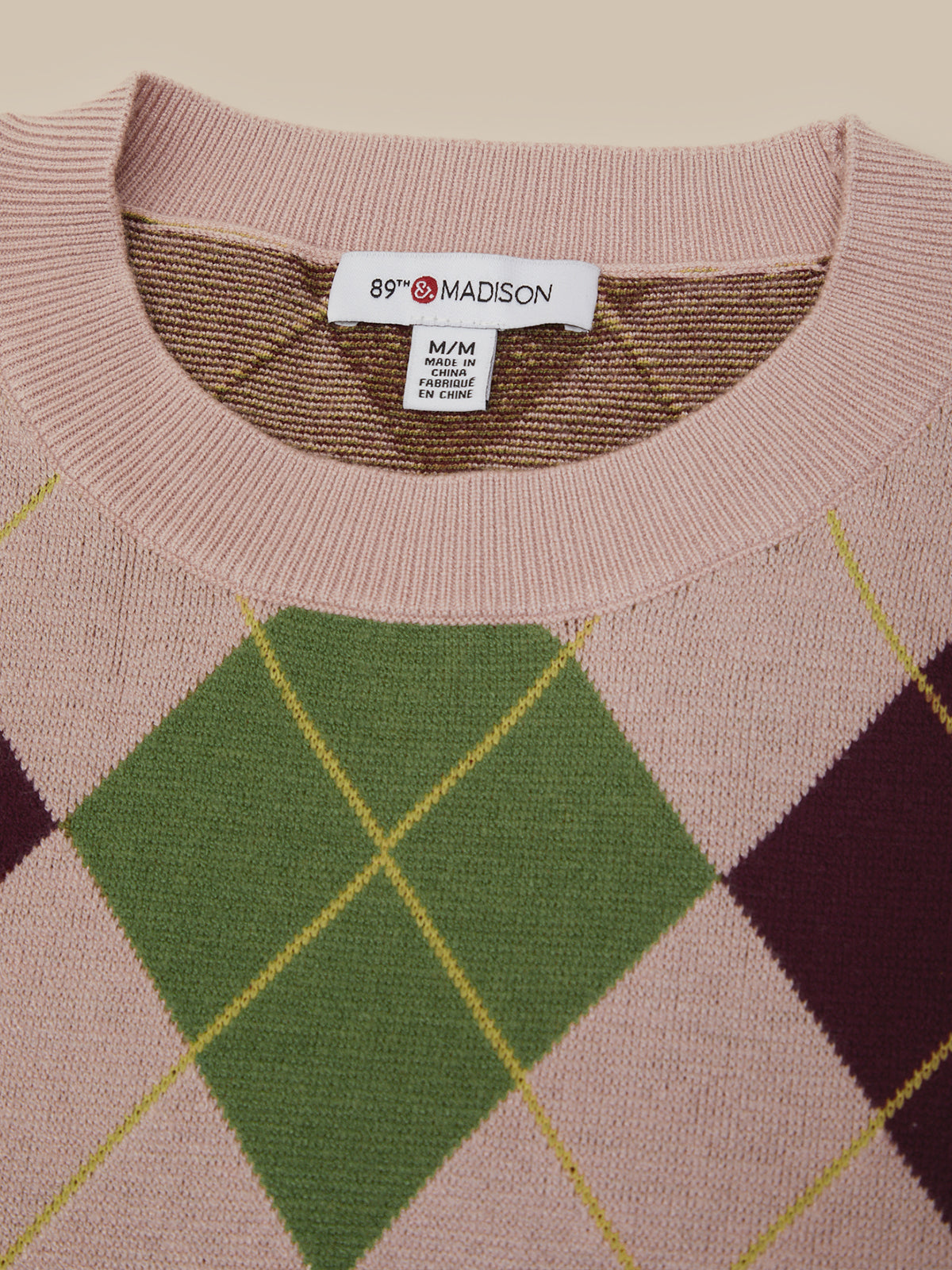 Close-up of a knit sweater with a dusty pink base and an argyle pattern featuring green, dark purple, and yellow diamonds. The brand label '89° MADISON' is visible inside the collar.