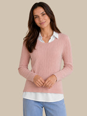 A woman wearing a pink ribbed knit sweater layered over a white collared shirt and light blue jeans, standing against a solid beige background.