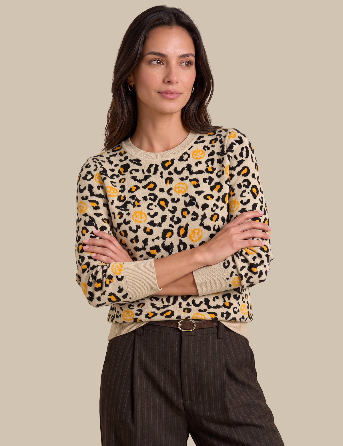 A woman modeling a beige crewneck sweater with a black and yellow leopard print pattern, paired with dark pinstriped trousers, against a solid beige background.