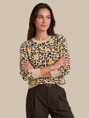 A woman modeling a beige crewneck sweater with a black and yellow leopard print pattern, paired with dark pinstriped trousers, against a solid beige background.