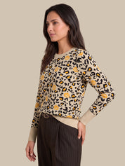 A woman modeling a beige crewneck sweater with a black leopard print and yellow floral accents, paired with dark brown pinstripe trousers, against a solid beige background.