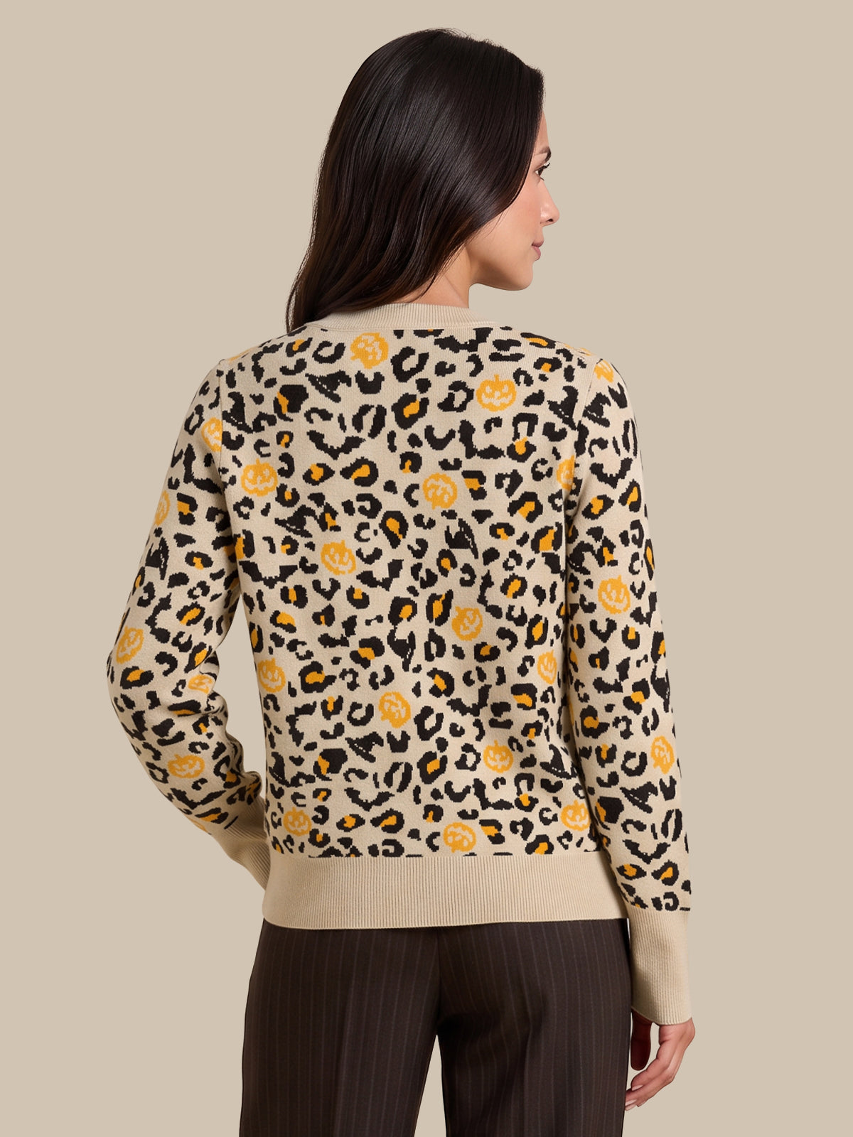 A woman shown from the back wearing a beige knit sweater with a black leopard print and yellow pumpkin motifs, paired with dark brown pinstripe pants, against a solid beige background.