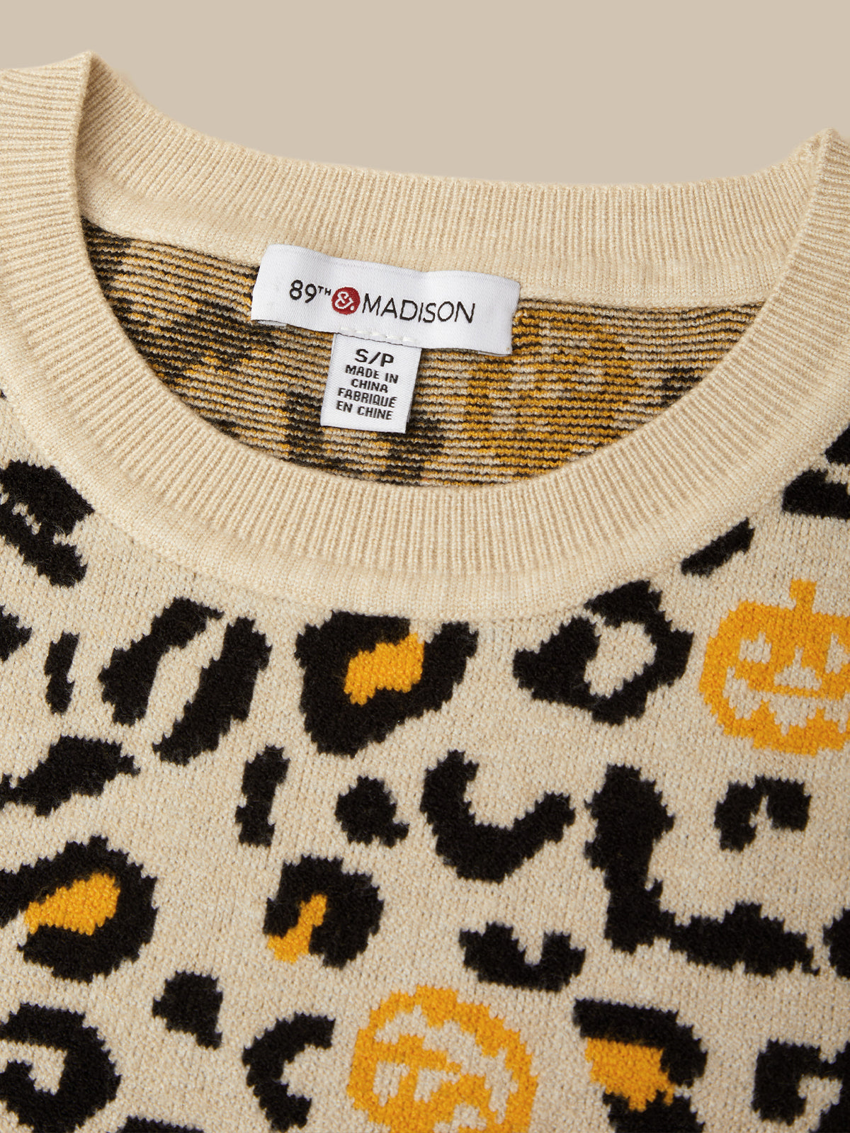 Close-up of a cream knit sweater with a black leopard print and yellow pumpkin motifs. The inside collar shows a white tag reading 89th Madison.