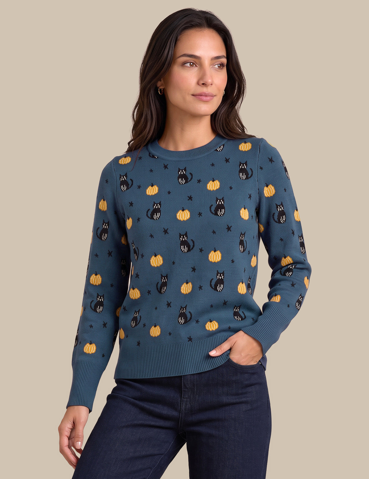A woman modeling a long-sleeve crewneck sweater in a slate blue color, decorated with a repeating pattern of black cats, orange pumpkins, and small black stars, paired with dark blue jeans against a solid beige background.