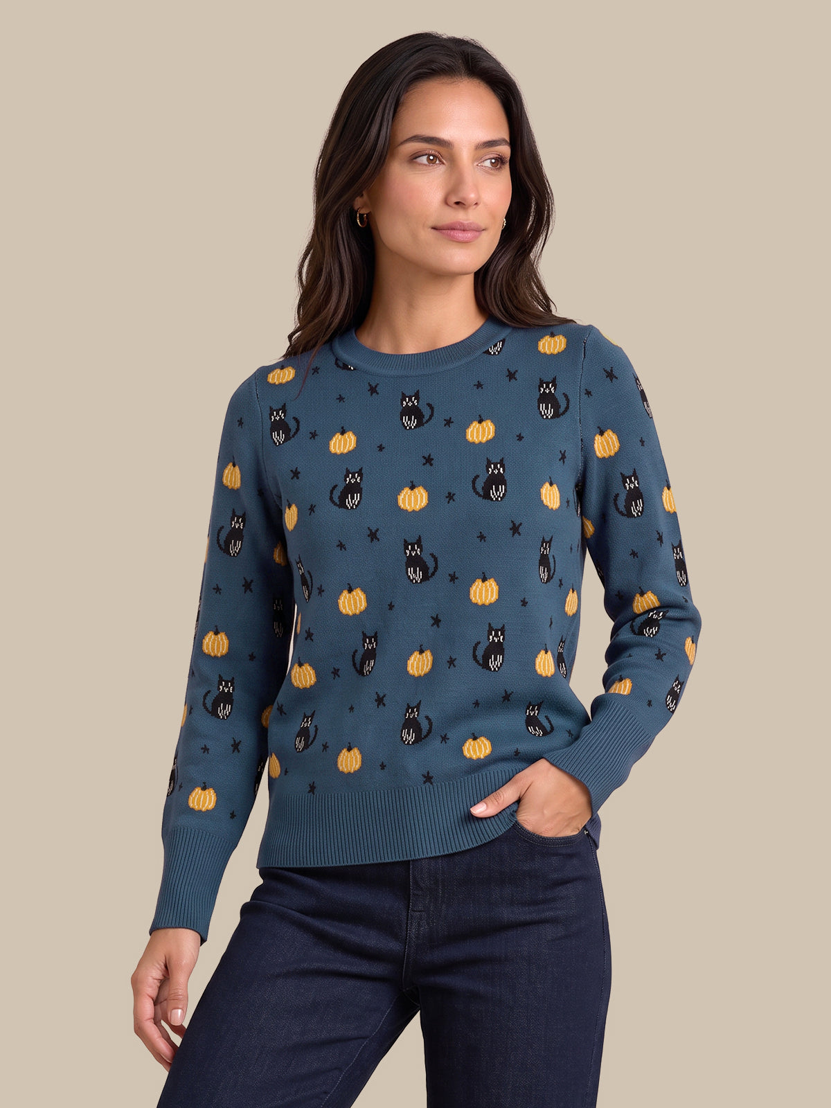 A woman modeling a long-sleeve crewneck sweater in a slate blue color, decorated with a repeating pattern of black cats, orange pumpkins, and small black stars, paired with dark blue jeans against a solid beige background.