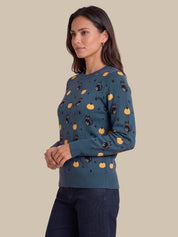 A woman modeling a long-sleeve crewneck sweater in a muted teal blue, decorated with a repeating pattern of black cats, orange pumpkins, and small black stars. She is paired with dark blue jeans against a solid beige background.