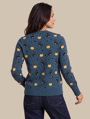 A woman shown from the back wearing a teal blue knit sweater with a repeating pattern of black cats, yellow pumpkins, and small black stars, paired with dark blue jeans against a solid beige background.