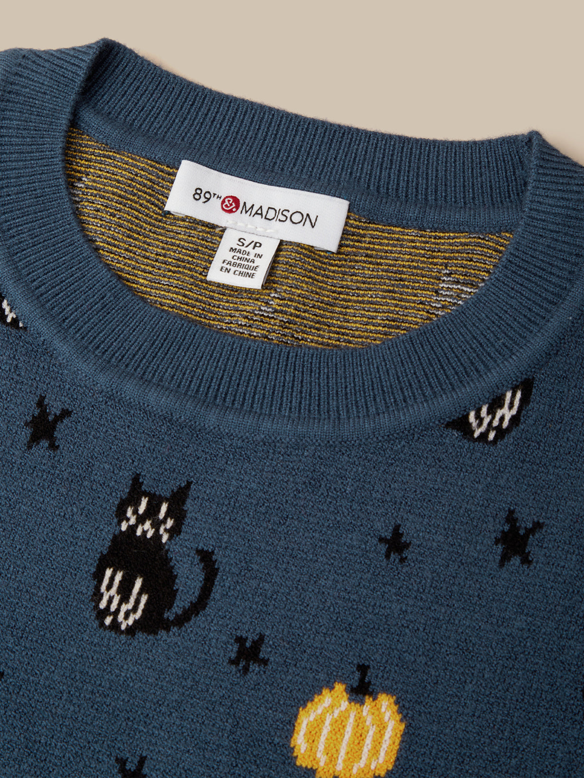 Close-up of a blue knit sweater with a crew neck, featuring a white 89th & Madison brand label inside the collar. The sweater has a pattern of black cats, yellow pumpkins, and black stars on a beige background.