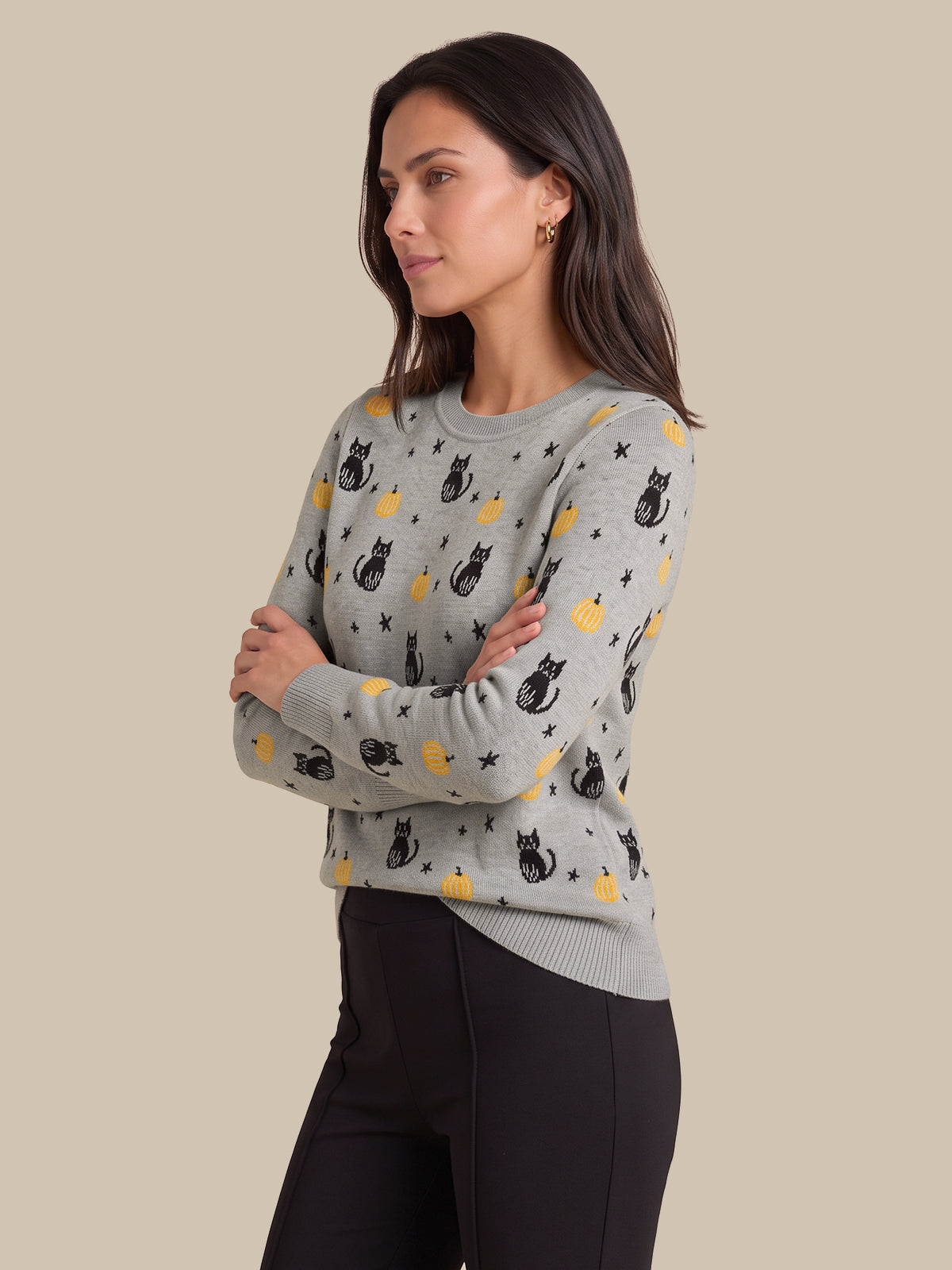 A woman modeling a light gray crewneck sweater with a pattern of black cats, yellow moons, and stars, paired with black pants, set against a solid beige background.