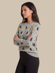A woman modeling a light gray crewneck sweater with a pattern of black cats, yellow moons, and stars, paired with black pants, set against a solid beige background.