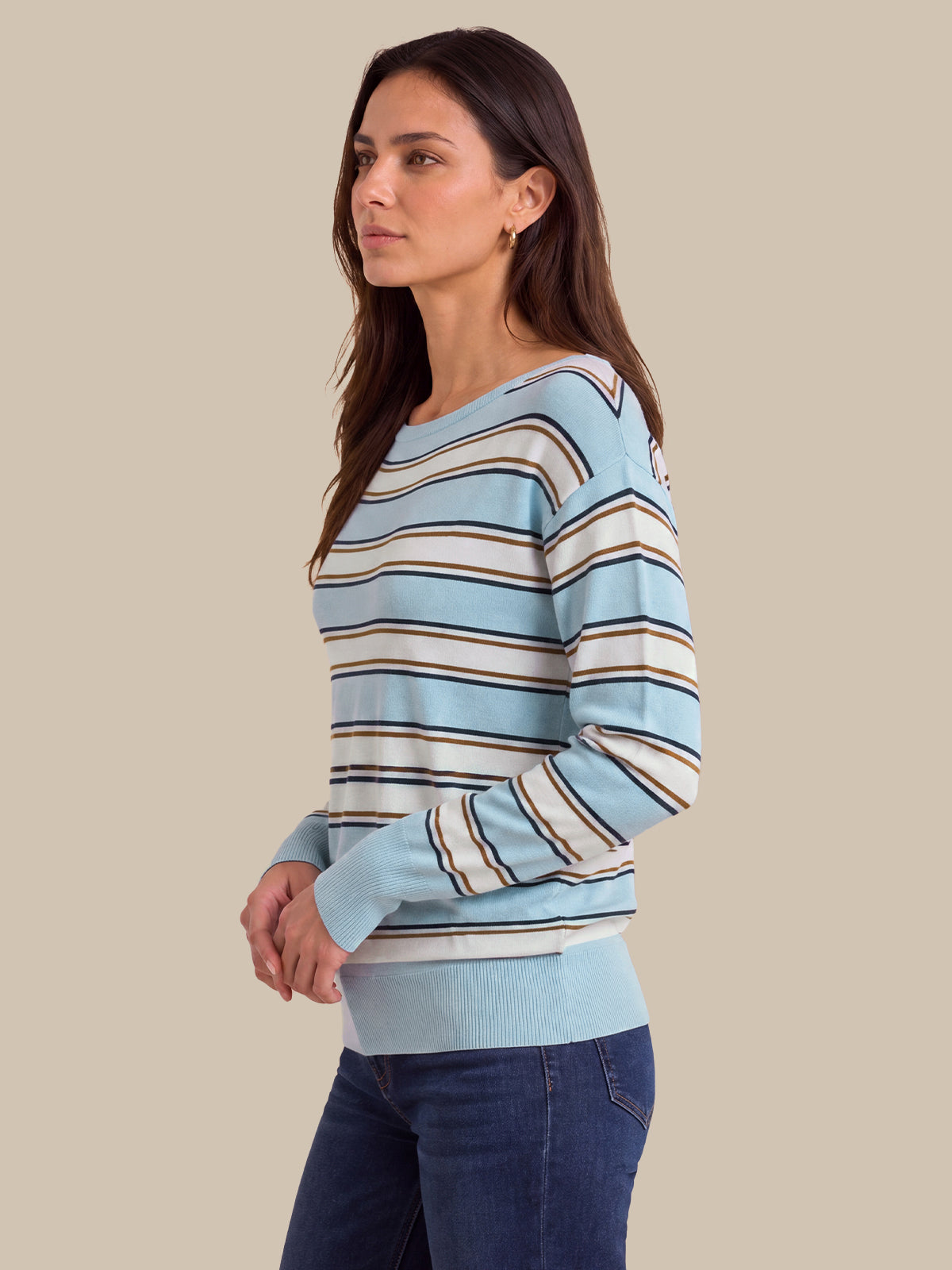 A woman wearing a light blue, white, and dark brown striped long-sleeve knit sweater, paired with dark blue jeans, shown from a side profile against a solid beige background.