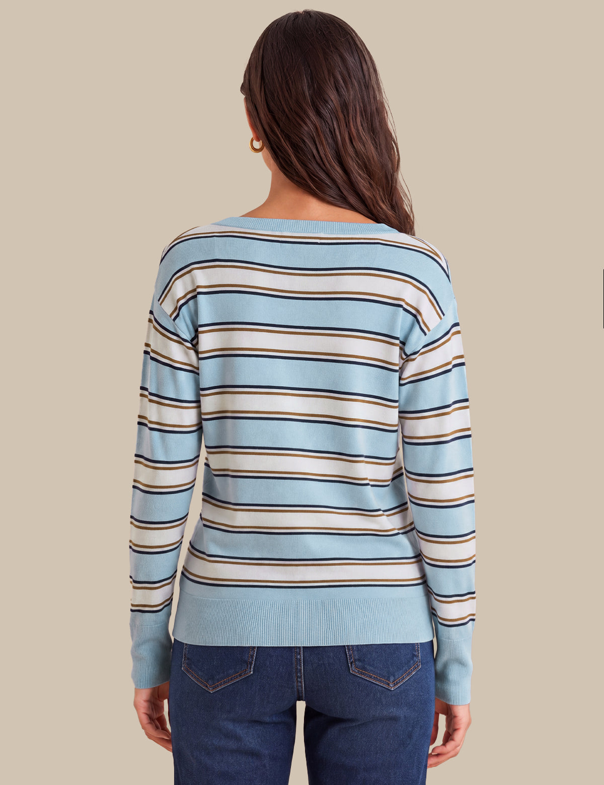 A woman viewed from the back wearing a light blue long-sleeve knit sweater with horizontal beige and dark brown stripes, paired with dark blue jeans, against a solid beige background.