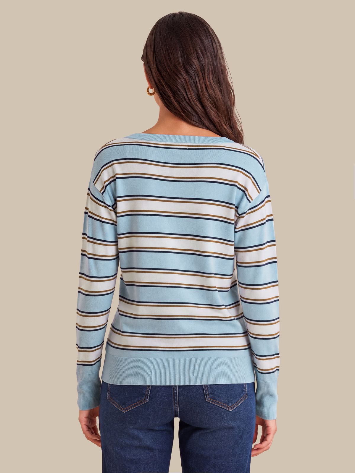 A woman viewed from the back wearing a light blue long-sleeve knit sweater with horizontal beige and dark brown stripes, paired with dark blue jeans, against a solid beige background.