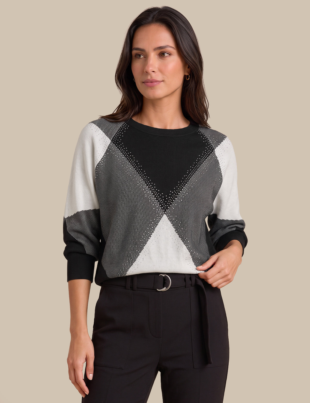 A woman modeling a long-sleeve knit sweater with a geometric color-blocked pattern in black, gray, and off-white, paired with black trousers against a solid beige background.