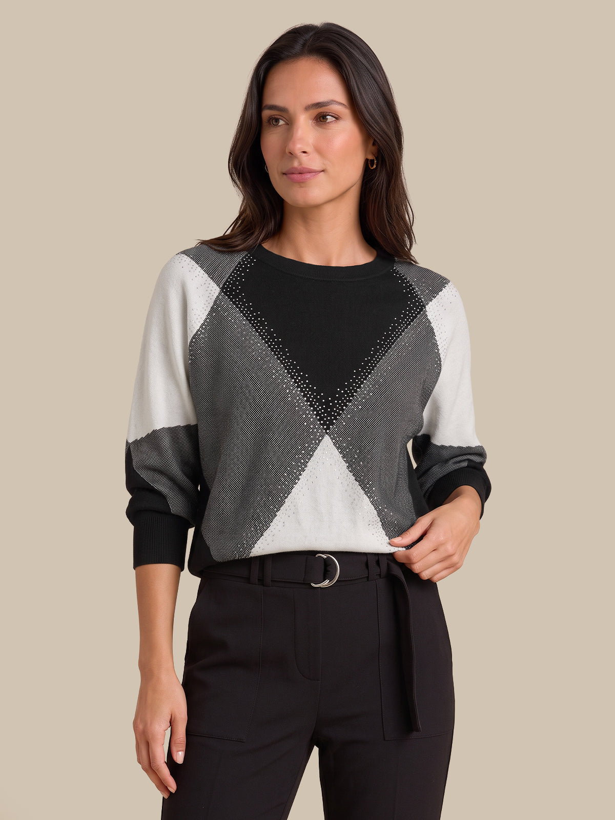 A woman modeling a long-sleeve knit sweater with a geometric color-blocked pattern in black, gray, and off-white, paired with black trousers against a solid beige background.