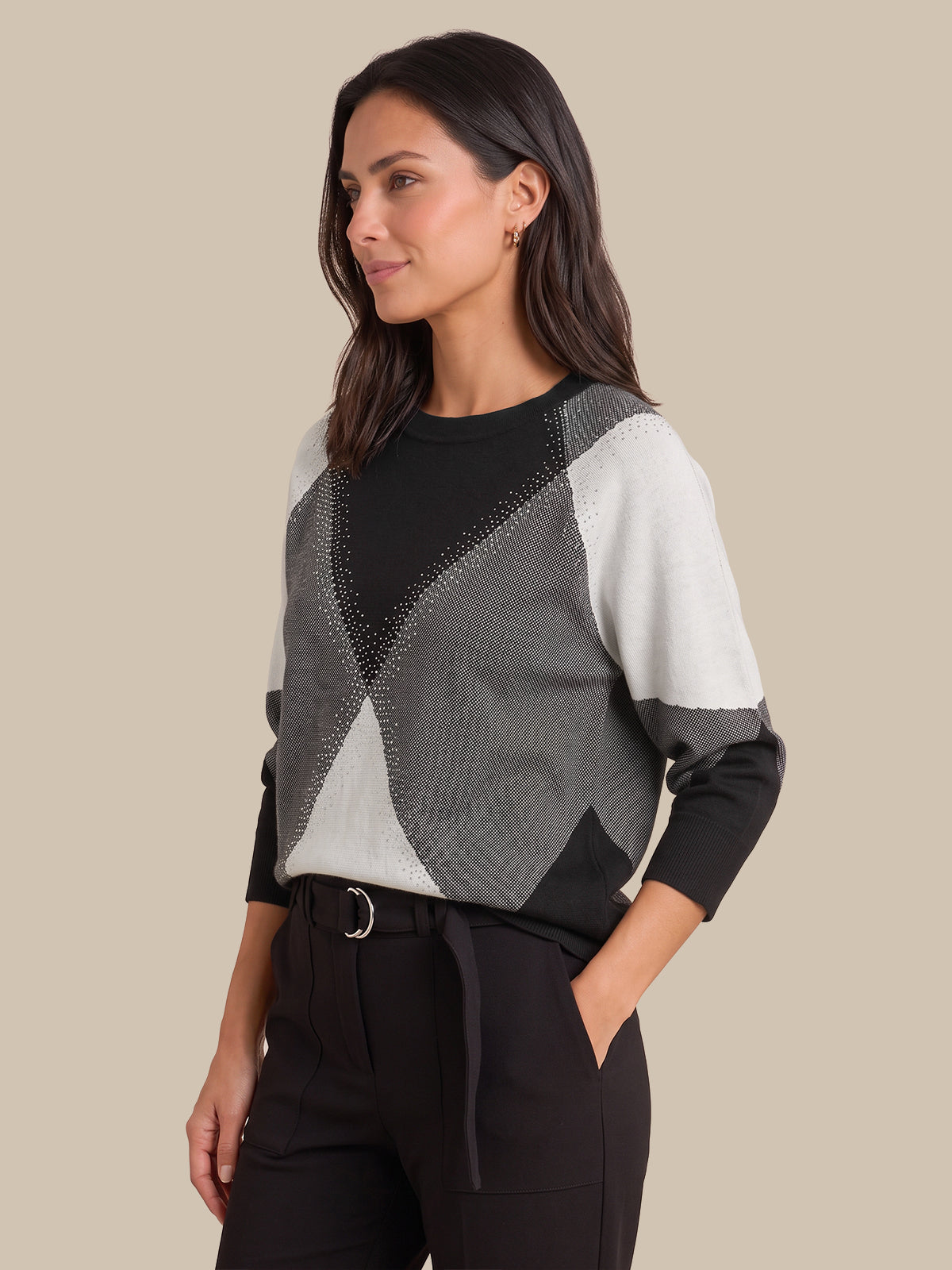 A woman modeling a color-block knit sweater with textured black, gray, and off-white panels, paired with black trousers, against a solid beige background.