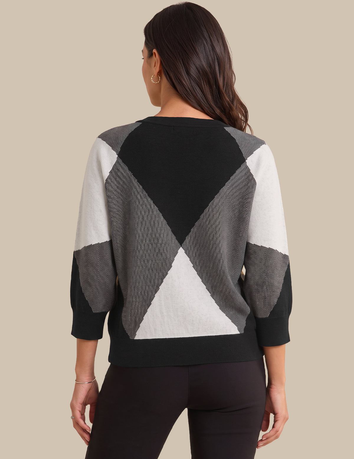 A woman viewed from the back wearing a geometric color-block knit sweater with black, gray, and white panels, paired with black leggings, against a solid beige background.