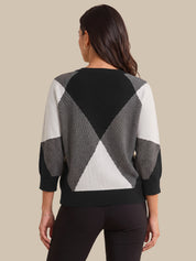A woman viewed from the back wearing a geometric color-block knit sweater with black, gray, and white panels, paired with black leggings, against a solid beige background.
