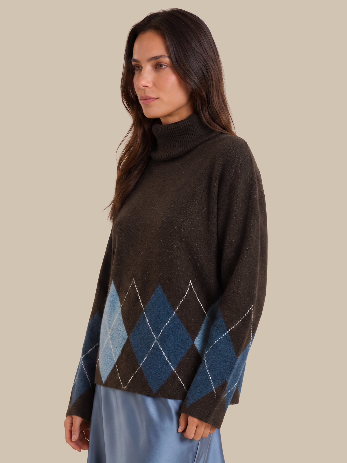 A woman wearing a dark brown turtleneck sweater with a blue and gray argyle pattern on the lower front and sleeves, paired with a light blue skirt, set against a solid beige background.