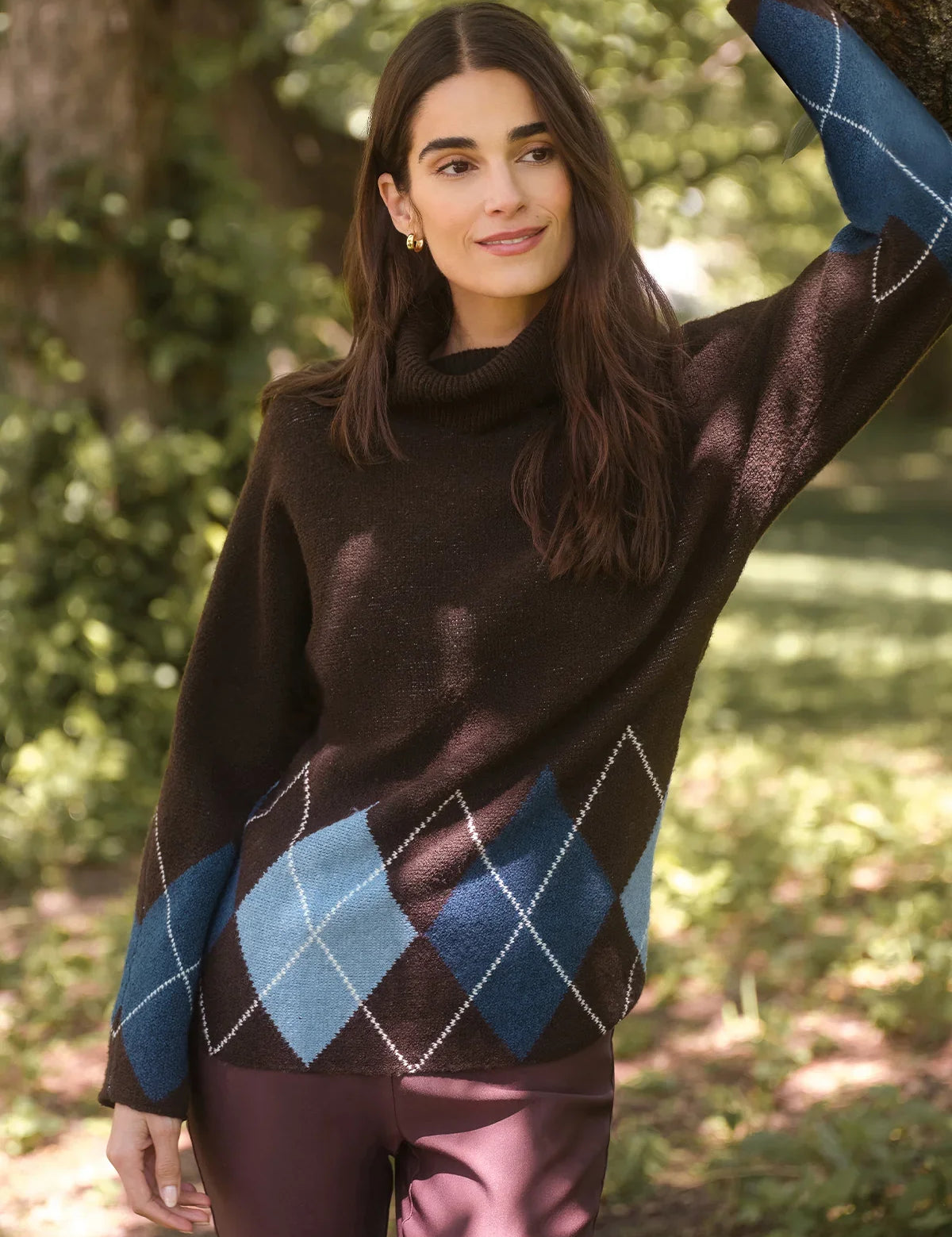 A woman wearing a dark brown turtleneck sweater with a blue and light blue argyle pattern on the lower half, paired with mauve pants, standing outdoors in a wooded setting.