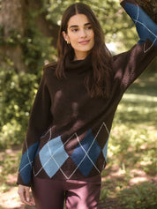 A woman wearing a dark brown turtleneck sweater with a blue and light blue argyle pattern on the lower half, paired with mauve pants, standing outdoors in a wooded setting.