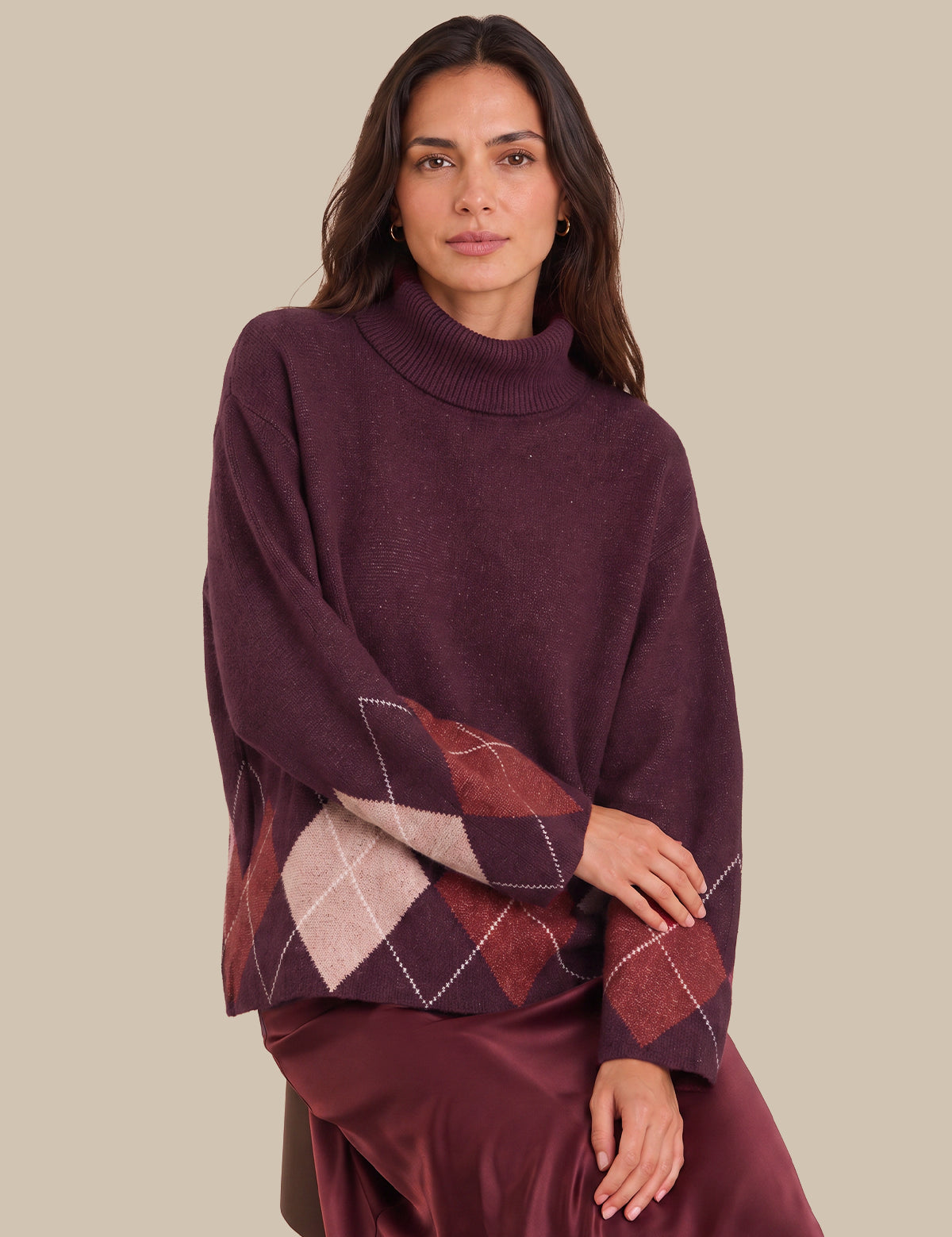 A woman wearing a dark plum purple turtleneck sweater with a contrasting argyle patterned sleeve, paired with a matching satin skirt, against a solid beige background.
