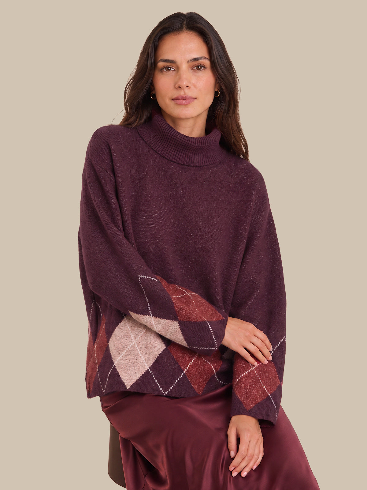 A woman wearing a dark plum purple turtleneck sweater with a contrasting argyle patterned sleeve, paired with a matching satin skirt, against a solid beige background.