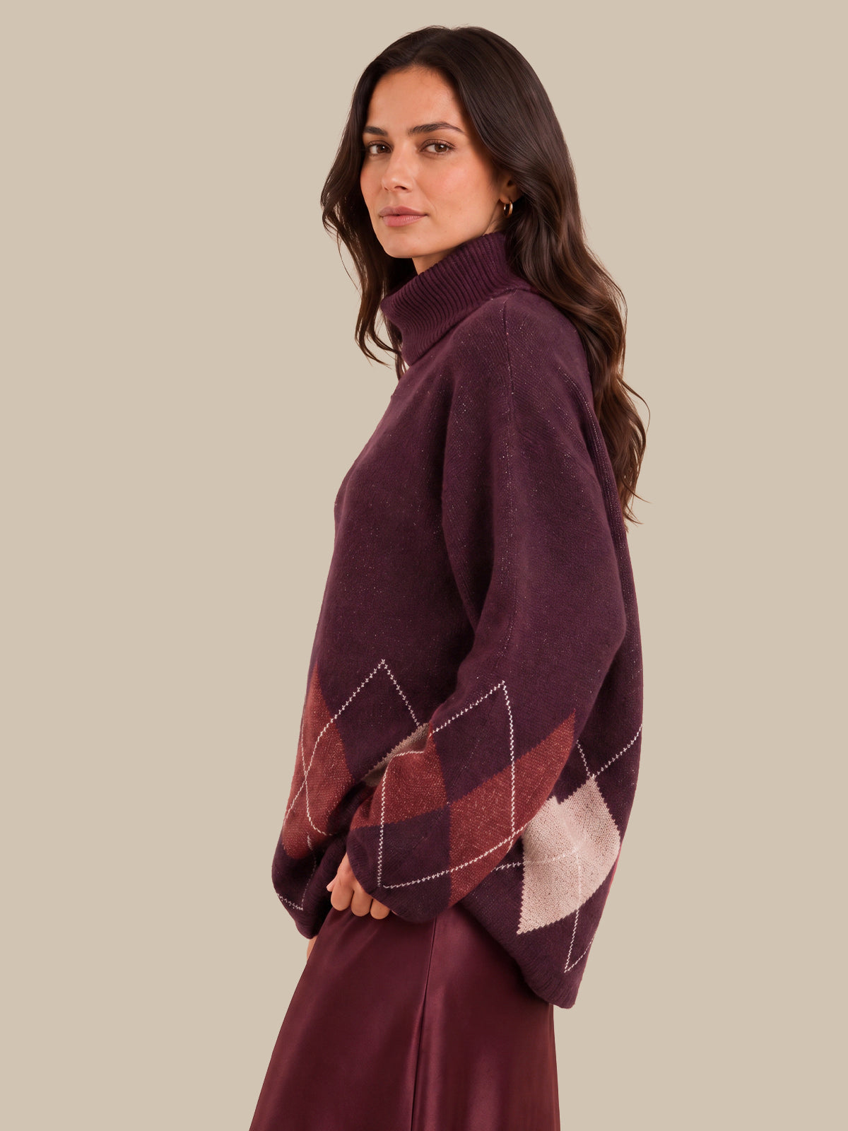 A woman wearing a dark plum purple knit sweater with argyle diamond patterns in burgundy and light beige on the lower sleeves, shown in a side profile against a solid beige background.
