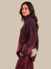 A woman wearing a dark plum purple knit sweater with argyle diamond patterns in burgundy and light beige on the lower sleeves, shown in a side profile against a solid beige background.
