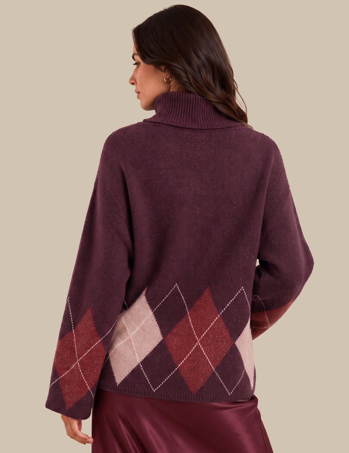 A woman shown from the back wearing a burgundy turtleneck sweater with an argyle diamond pattern in burgundy, dusty pink, and white on the lower back and sleeves, paired with a burgundy satin skirt against a solid beige background.
