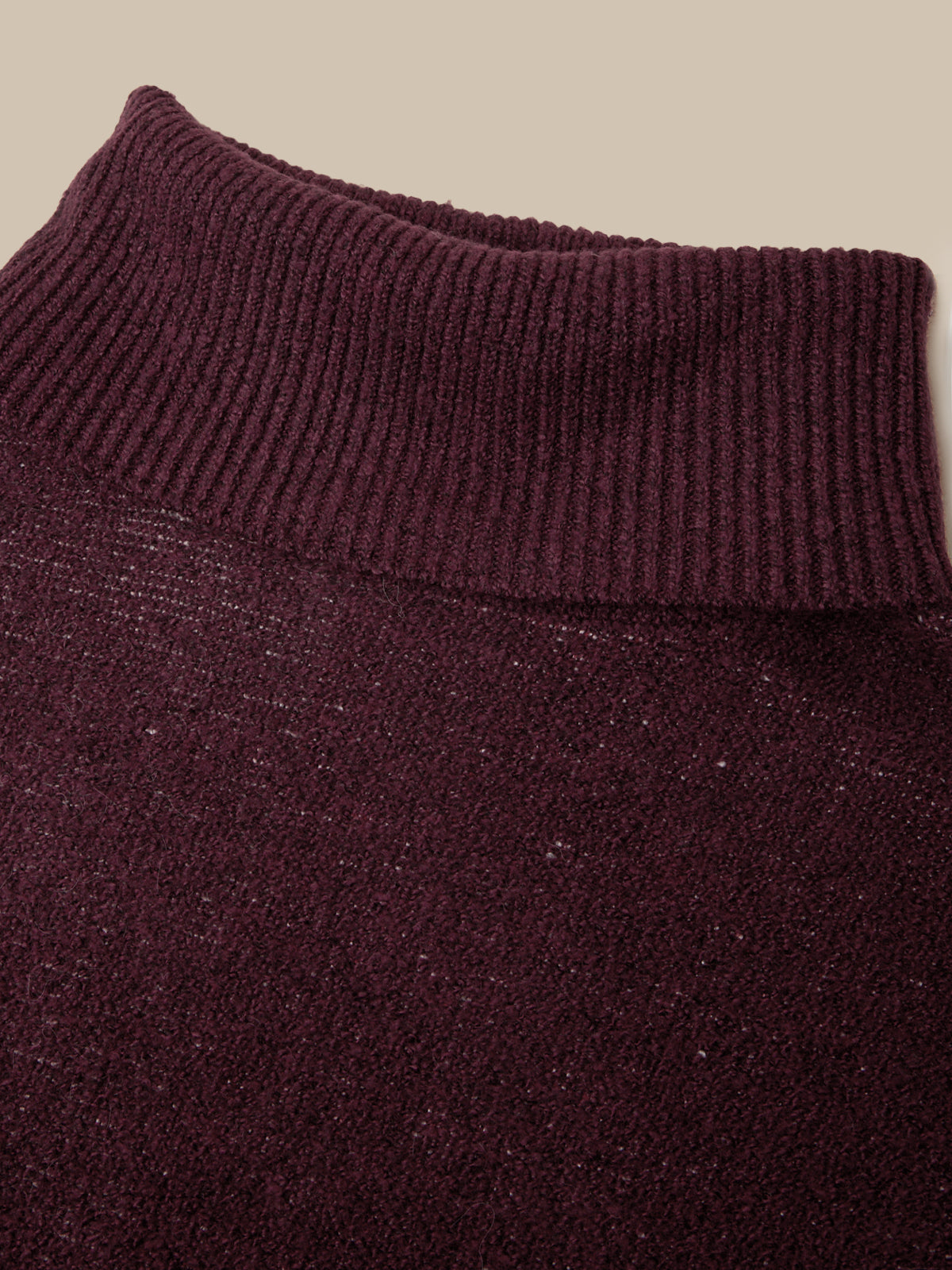 Close-up of a ribbed knit turtleneck sweater in a deep burgundy or plum purple color, showing the texture and ribbed collar against a solid beige background.