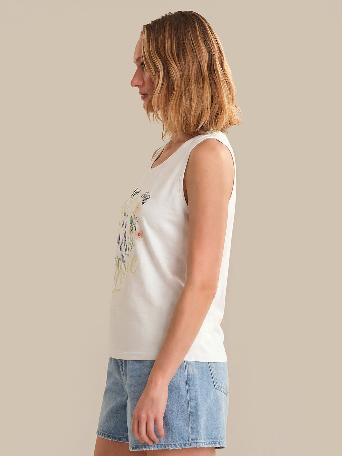 Woman wearing a white sleeveless top with floral design and denim shorts on a beige background