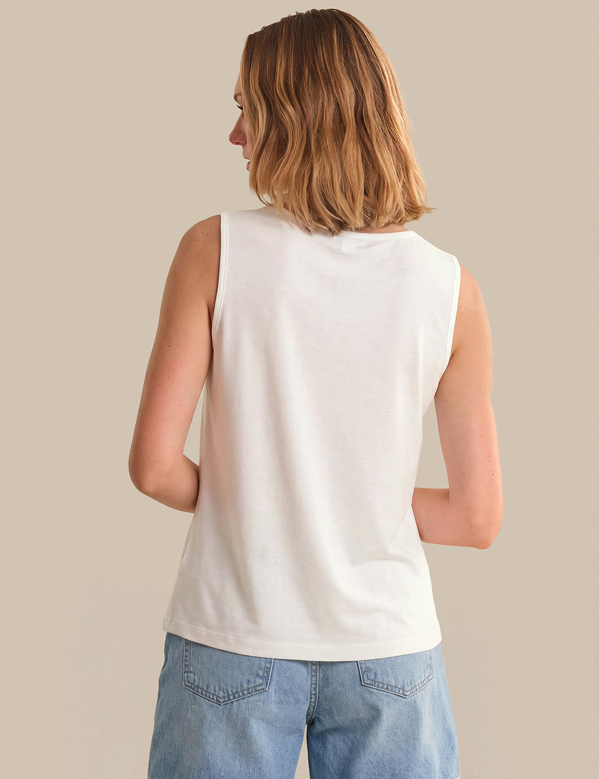 Woman wearing a white sleeveless top and blue jeans against a beige background