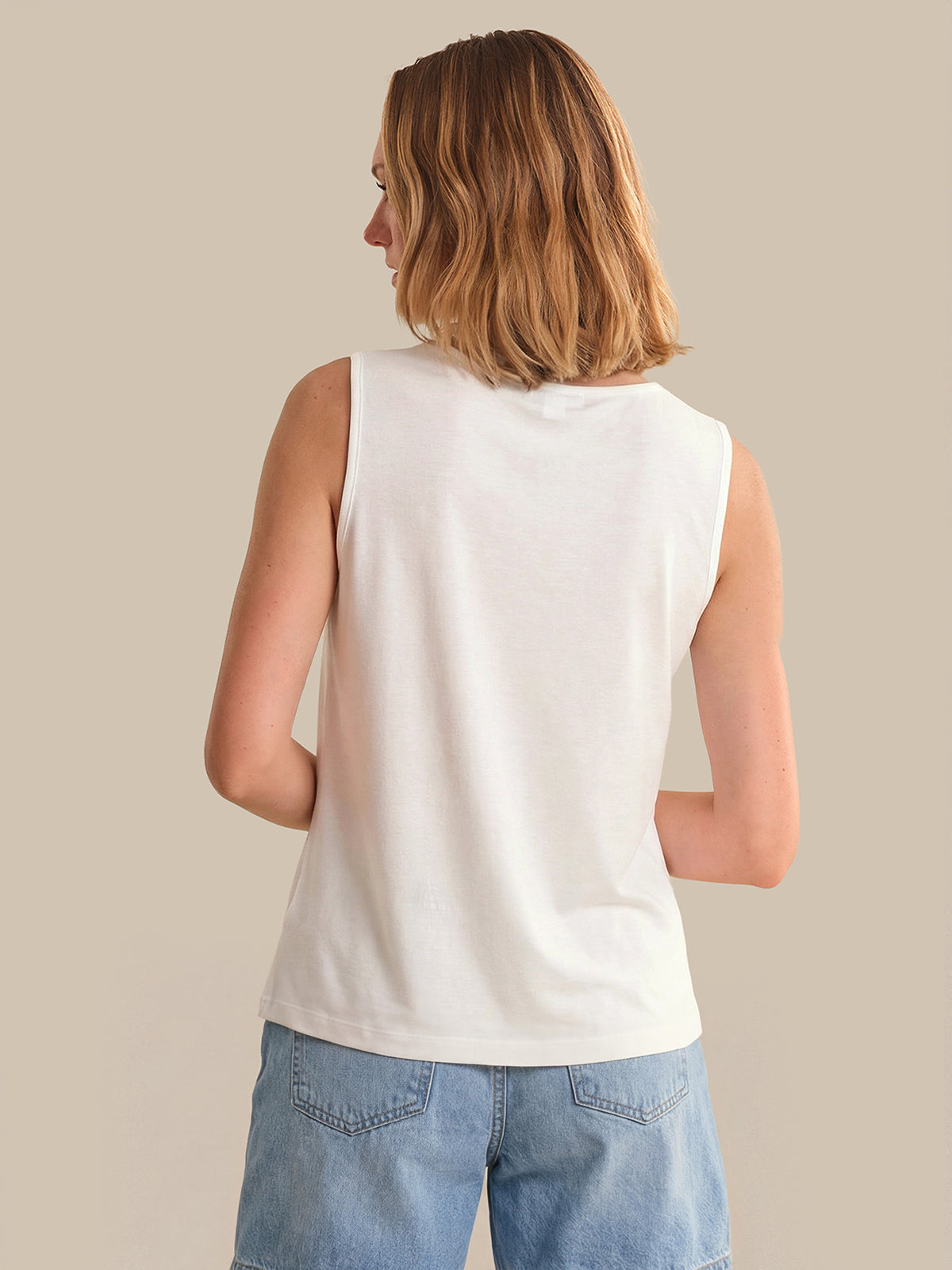 Woman wearing a white sleeveless top and blue jeans against a beige background