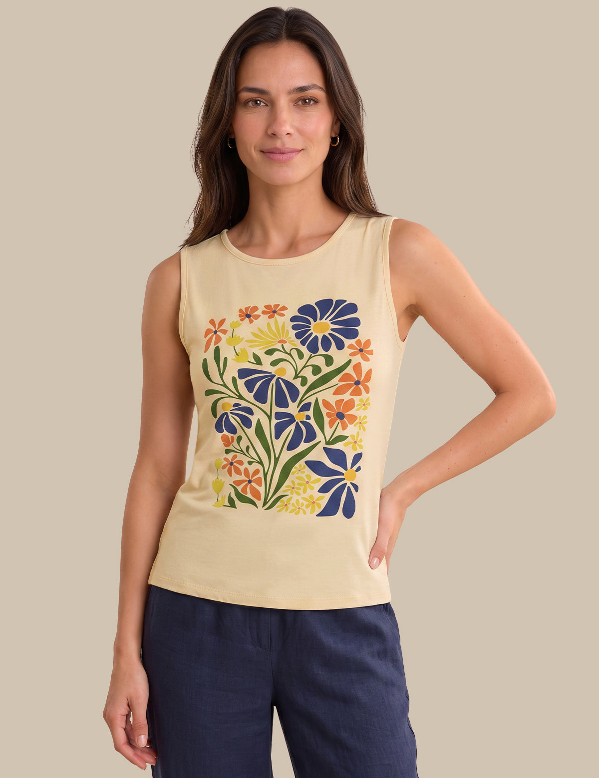 A woman modeling a sleeveless cream-colored tank top with a colorful floral print on the front, paired with navy blue pants, set against a solid beige background.